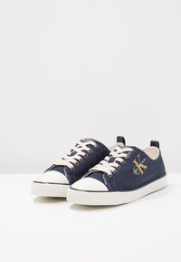Jeans sneakers med vit gummikappa och sula, krämfärgade snörning och en guldfärgad logotyp på sidan. Rundad form med låg profil design.