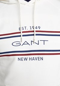 Felpa bianca con cappuccio e cordini, caratterizzata da strisce orizzontali blu e bordeaux. Sul davanti appare la scritta "GANT" e "NEW HAVEN" con "EST. 1949" sopra.