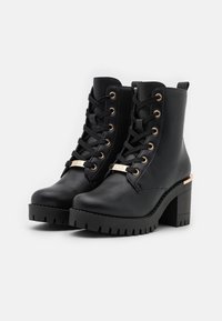 Bottines en cuir noir avec œillets dorés, laçage frontal, un talon épais et une semelle crantée. Plaque dorée en accent sur le côté.
