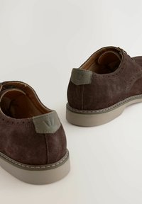 Braune Wildlederschuhe mit grauen Akzenten, runder Zehenform und einer hellgrauen Sohle. Besitzt durchbrochene Details und einen gestickten Rand.