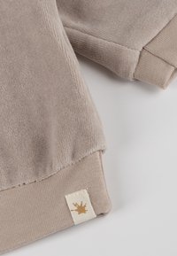 Beiger Pullover aus weichem Fleece-Material mit strukturierter Oberfläche. Verfügt über gerippte Bündchen und ein kleines Etikett mit braunem Farbspritzendesign.