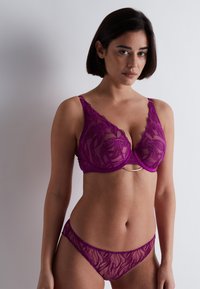 Soutien-gorge en dentelle violette avec des motifs floraux et un accent doré au centre. String en dentelle violette transparente assorti. Les deux ont une finition délicate et texturée.