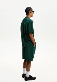 Zöld pamut póló és hozzá illő rövidnadrág, laza szabás, oldalsó zsebekkel, fekete sportcipővel és fehér, térd alatti zoknival viselve. Egyszerű design, minta nélkül.