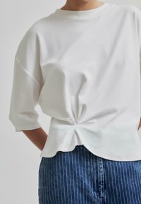 Balta medvilninė marškinė su apvaliu kaklu ir trijų ketvirčių rankovėmis, su surištu akcentu nugaros dalyje, derinami su mėlynomis dryžuotomis džinsinėmis kelnėmis.