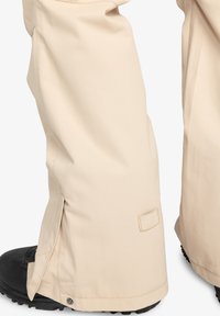 Beige snow pants with a loose fit, featuring side zippers, a patch pocket, and reinforced stitching, paired with black snow boots.