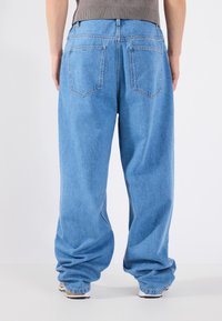 Jeans de ganga azul claro com um corte descontraído, apresentando dois bolsos traseiros e um ligeiro elastano no tecido, usados com um suéter cinza.