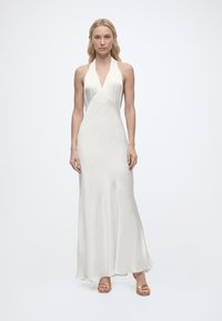 Γυναίκα που φοράει λευκό σατέν φόρεμα χωρίς μανίκια με λαιμόκοψη halter και χρυσά ανοιχτά πέδιλα, στέκεται μπροστά σε απλό λευκό φόντο.