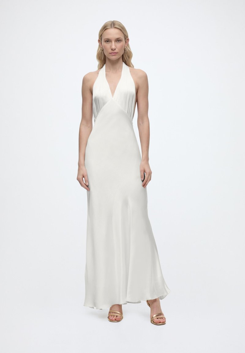 Γυναίκα που φοράει λευκό σατέν φόρεμα χωρίς μανίκια με λαιμόκοψη halter και χρυσά ανοιχτά πέδιλα, στέκεται μπροστά σε απλό λευκό φόντο.