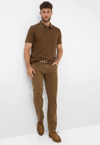 Polo shirt marrone con leggere strisce sulle maniche, abbinato a pantaloni marroni e scarpe in suede marroni. Il modello è in piedi contro uno sfondo bianco semplice.