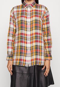 Person wearing a multicolored plaid button-up shirt with long sleeves and a black leather skirt, standing against a plain background.