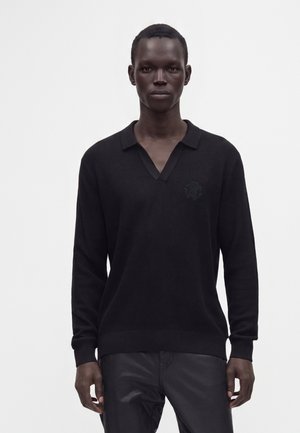 Homme portant une chemise en tricot noire à manches longues avec col et un pantalon noir, debout devant un fond blanc uni.