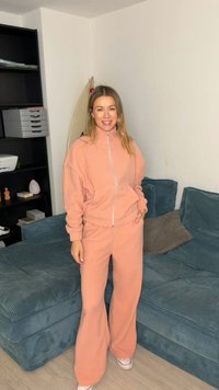 Korallenfarbener Fleece-Trainingsanzug mit Reißverschlussjacke und weiten Hosenbeinen. Weiche Textur, lockere Passform und abgestimmtes Farbschema. Weiße Sneaker.