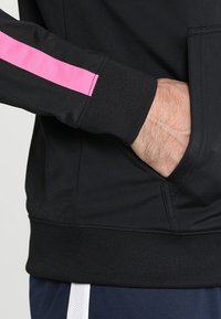 Schwarze Sportjacke mit strukturiertem Finish, ausgestattet mit einem pinken Streifen am Ärmel und einer in das Design integrierten Fronttasche.