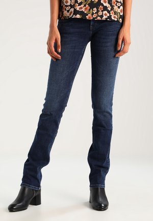 Vrouw met donkerblauwe slim-fit jeans, een bloemen top en zwarte enkelhoge hakken, staand met licht gespreide benen tegen een lichte achtergrond.