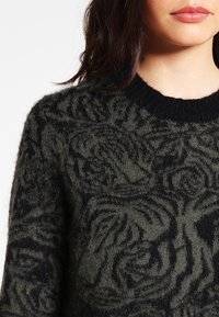 Dziany sweter w ciemnozielonym kolorze z fakturowanym wzorem czarnej róży. Posiada okrągły dekolt i miękki, puszysty materiał.