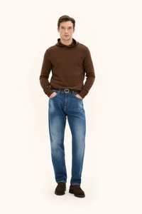 Brauner gestrickter Hoodie, blaue gerade Jeans, braune Wildlederschuhe, Hände in den Taschen, neutraler Hintergrund.