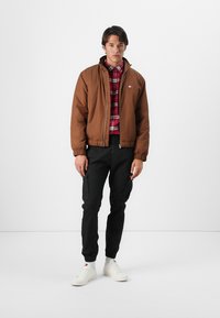 Brun zip-up bomberjakke med ribbede manchetter, båret over en rød ternet skjorte og parret med sorte cargo bukser og hvide sneakers.