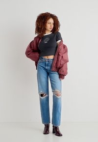 Γυναίκα με σγουρά μαλλιά που φοράει μαύρο κοντό τοπ, μπορντό μπουφάν που έχει κατεβασμένα τους ώμους, σκισμένο μπλε τζιν και σκούρα μποτάκια μέχρι τον αστράγαλο, στέκεται μπροστά σε λευκό φόντο.