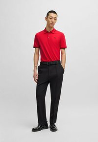 Rød polo skjorte med sorte accenter og logo, parret med sorte bukser og sorte sko, stående mod en lys grå baggrund.