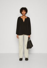 Svart langermet bluse med V-hals og samlet skuldrer, kombinert med lys beige vide bukser og en liten svart toalettveske.