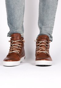 Sneaker alte in pelle marrone con suola in gomma bianca, occhielli metallici e lacci beige. Superficie testurizzata e logo sulla linguetta.