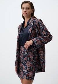 Ensemble blazer et jupe en velours marine avec des motifs paisley complexes en rouge, bleu et vert. Tissu lisse avec une coupe ajustée.