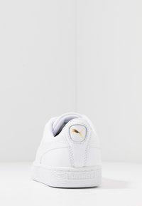 Bílé tenisky s hladkým koženým povrchem, polstrovaným límcem a texturovanou gumovou podešví. Na zadní části paty je zlatý logo akcent.