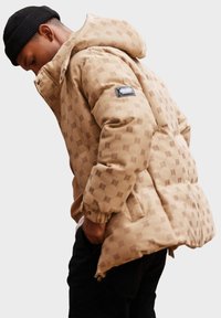 Béžová prošívaná bunda s kapucí, která má jemný čtvercový vzor, elastické manžety a boční kapsu s výrazným logem.