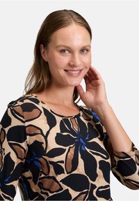 Długi rękaw bluzka z kwiatowym wzorem w kolorze czarnym, niebieskim i brązowym na beżowym tle; okrągły dekolt i miękka tkanina.