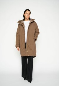 Parka imbottita marrone con cappuccio, dotata di chiusura con zip e bottoni a pressione, tasche laterali e una fodera interna liscia. Indossata sopra pantaloni larghi scuri.