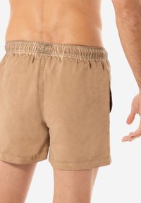 Pantaloni da bagno marroni con una vita elasticizzata, tessuto testurizzato e tasche laterali, caratterizzati da cuciture minime e un taglio classico.