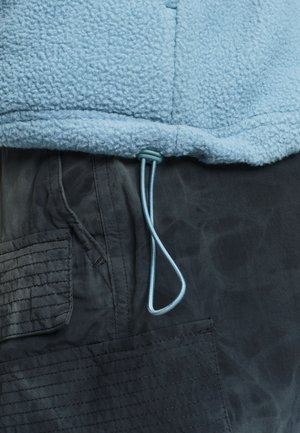 Ourlet de veste en polaire bleu clair avec cordon de serrage réglable sur une poche de pantalon cargo gris foncé avec des détails cousus.