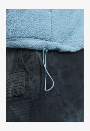 Lichtblauwe fleecejackzoom met verstelbare trekkoord over zak van donkergrijze cargobroek met gestikte details.