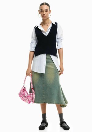 Nő réteges, ujjatlan fekete pulóverben csíkos ing fölött, farmer midi szoknyában, szürke zokniban, fekete cipővel, és rózsaszín táskát tartva.