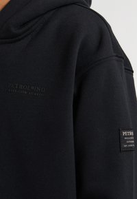Sweat à capuche noir en tissu doux, avec un logo texturé imprimé et une étiquette sur la manche pour des fins de marquage.
