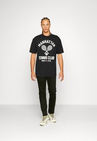 Juodas medvilninis marškinėlis su baltu "Manhattan Tennis Club" užrašu ir teniso raketės grafika, derinamas su tamsiai žaliais kelnių ir baltais sportbačiais.