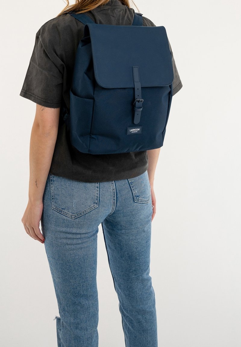 Personne portant un t-shirt sombre et un jean bleu avec un sac à dos carré bleu marine avec rabat et fermeture à boucle dans le dos.