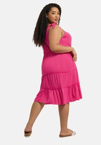 Rosa lagerklänning med justerbara knytband, lätt textur och utsvängd nederdel. Modellen bär platta sandaler med korkdetaljer.