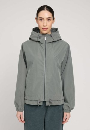 Woman wearing a muted green hooded jacket with front zipper and drawstrings, standing against a plain light background.