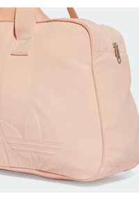 Růžovooranžová sportovní taška vyrobená z hladké tkaniny, s uzavíráním na zip a vyraženým logem se třemi pruhy na boku.