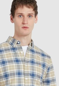 Een beige, blauwe, en witte geruite overhemd met een spread collar; heeft een klein geborduurd logo en standaardknopen.