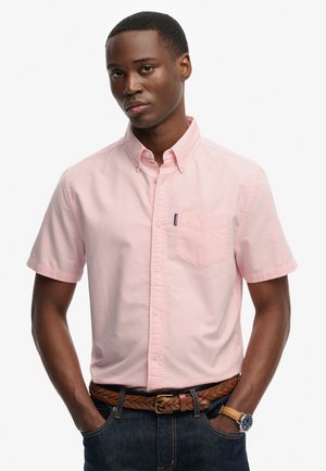 Mężczyzna w jasnoróżowej, krótkorękawowej koszuli na guziki, ciemnych dżinsach, brązowym plecionym pasku i zegarku z brązowym skórzanym paskiem.