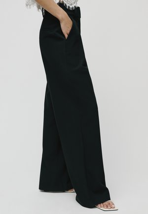 Zwarte wijde broek van gladde stof, met zijzakken en een hoge taille. Schoenen zijn open tenen met een minimalistische stijl.