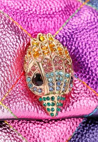 Gouden broche met kleurrijke edelstenen in een schelpdessin, gemonteerd op een gestructureerde roze en paarse leren achtergrond met kleurrijke stiksels.