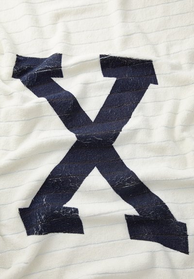 Biela osuška s textúrovaným povrchom, zdobená veľkým námořnicky modrým dizajnom "X". Pozadie má jemné pruhované vzory.