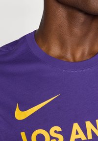 Violetinės medvilnės marškinėliai su geltonu Nike swoosh ir ryškiai geltonu užrašu "LOS ANGELES". Apvalus kaklas ir glotni tekstūra.
