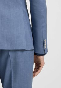 Niebieska dopasowana marynarka o gładkiej fakturze. Posiada jedno rozcięcie z tyłu oraz cztery jasne guziki na mankiecie.