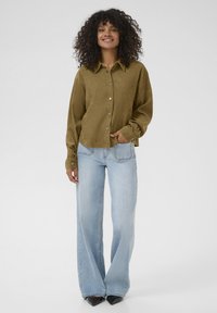Olijfgroene corduroy blouse met knoopsluiting, gecombineerd met lichtblauwe wijde jeans en zwarte puntige hakken. Gladde texturen zichtbaar.