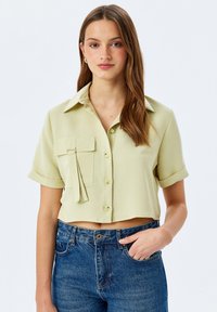 Lichtgroen cropped shirt met korte mouwen, een kraag, een zak aan de voorkant en knoopsluiting. Model gecombineerd met blauwe denim jeans.