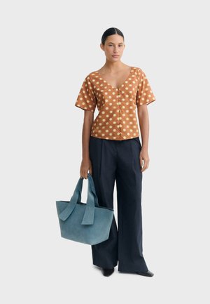 Femme portant un chemisier marron à manches courtes à pois, un pantalon large noir, des chaussures noires, tenant un grand sac cabas bleu à côté d'elle.